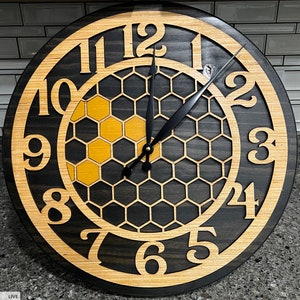 Può includere: Un orologio nero e dorato con un design a nido d'ape. Il quadrante dell'orologio ha numeri romani e lancette nere. L'orologio è in legno e ha una finitura invecchiata.