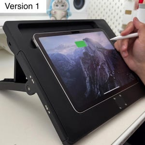 May include: A black tablet stand with an adjustable angle, holding a tablet displaying a mountain landscape. A white stylus is touching the screen. The tablet shows a green charging indicator. The stand is on a white surface.