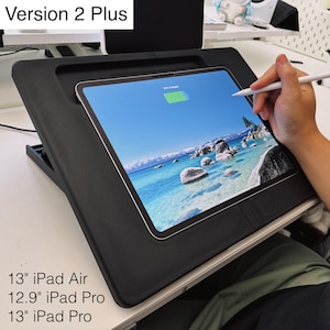 May include: A black angled tablet stand labeled Version 2 Plus, compatible with 13 inch iPad Air, 12.9 inch iPad Pro, and 13 inch iPad Pro. The stand holds a tablet displaying a landscape image of a blue lake with rocks, and a white stylus is in use.
