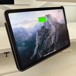 May include: A black tablet with a landscape image of a mountain range on the screen. The tablet is charging, indicated by a green battery icon showing 80% charged. The tablet is in a black case.