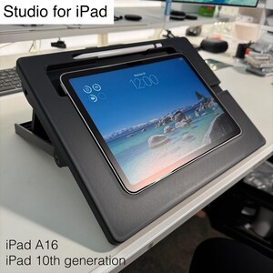 May include: A black tablet stand labeled Studio for iPad, holding an iPad displaying a beach scene with the time 12:00. The stand features a dedicated slot for a white stylus at the top. Text on the image reads Studio for iPad, iPad A16, and iPad 10th generation.