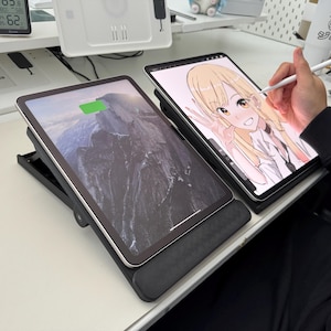 May include: Two tablets are displayed on black stands. One tablet shows a mountain landscape, and the other displays a digital drawing of an anime character. A stylus is held near the second tablet.