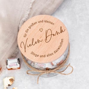 Könnte beinhalten: Ein klares Glas mit einem Holzdeckel, mit Bindfaden gebunden. Der Deckel ist mit dem deutschen Text "Vielen Dank" graviert. Das Glas ist mit verpackten Süßigkeiten gefüllt. Einige Süßigkeiten sind außerhalb des Glases sichtbar.