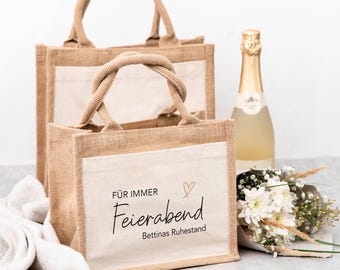 May include: Two beige burlap tote bags with cream-colored panels and twisted rope handles. The front bag has the words "FÜR IMMER Feierabend Bettinas Ruhestand" printed on it. A bottle of champagne and a bouquet of white flowers are next to the bags.