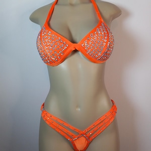 Può includere: Bikini arancione con strass argentati. La parte superiore è di stile halter con un reggiseno push-up. La parte inferiore è di stile a taglio alto con un design incrociato.