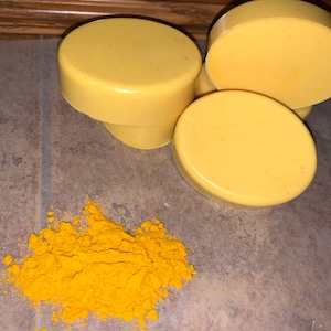 May include: A collection of light yellow, oval-shaped soaps alongside a pile of bright yellow powder. The soaps have a smooth texture. The powder is finely ground, creating a vivid contrast with the soaps. The scene suggests a natural skincare product.