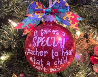 May include: A round red glittery Christmas bauble featuring white text that reads, It takes a SPECIAL teacher to hear what a child can't say. The bauble is topped with a colourful bow featuring a multi-coloured puzzle piece pattern.