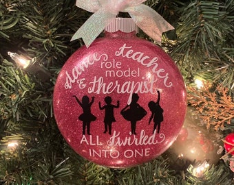 Puede incluir: Una bola de Navidad rosa brillante con texto blanco que dice "dance teacher role model & therapist ALL twirled INTO ONE." Cuatro siluetas negras de bailarines están debajo del texto. La bola está rematada con un lazo blanco.
