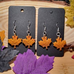 Puede incluir: Un par de pendientes colgantes con un diseño de hoja de arce. Las hojas están hechas de arcilla polimérica naranja y amarilla y están unidas a ganchos plateados.