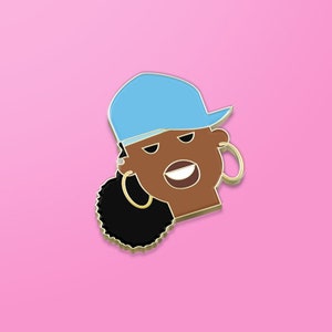 May include: A gold-colored enamel pin featuring a cartoon portrait of a person with brown skin. The person is wearing a light blue baseball cap and large gold hoop earrings. The person has black hair pulled back in a ponytail.