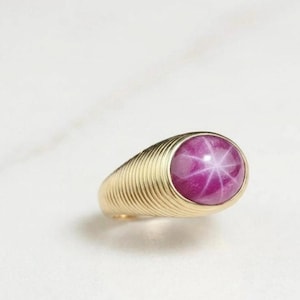 May include: A gold ring with a large, oval star ruby gemstone. The ruby is a deep pink colour and displays a six-rayed star pattern. The ring's band has a ridged design.