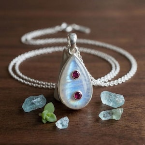 May include: A silver necklace featuring a teardrop-shaped pendant with a large, iridescent moonstone cabochon. Two small, round, deep pink rubies are bezel-set vertically along the center of the moonstone. The pendant hangs from a delicate silver chain.