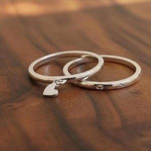 May include: Two silver-toned rings with a polished finish. One ring features a small, dangling heart-shaped charm attached to the band, while the other ring displays a series of small, engraved circular and cross-shaped symbols along its surface.
