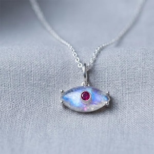 May include: A silver necklace with a pendant shaped like an eye. The pendant has a blue and white iridescent stone with a small red gemstone in the center. The necklace is on a gray fabric background.
