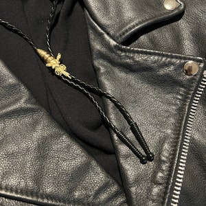 May include: A black leather jacket with a zipper closure and a bolo tie with a gold bee charm.