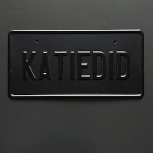 May include: A rectangular black metal license plate featuring the embossed text KATIEDID in capital letters. The plate has a raised border and two circular mounting holes at the top, presenting a solid matte finish with a clean, industrial design.