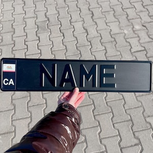 Puede incluir: Placa de matrícula personalizada negra con la palabra "NAME" y la abreviatura del estado "CA" en letras blancas. La placa tiene una pequeña bandera del estado de California en la esquina superior izquierda.