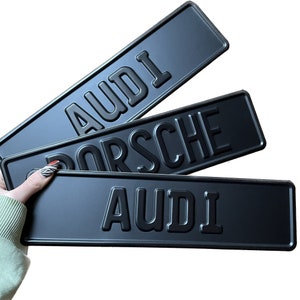 May include: Three black metal signs with raised lettering. The signs read "AUDI", "PORSCHE", and "AUDI".