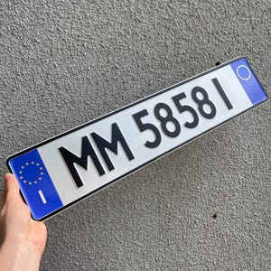 May include: A rectangular license plate with the text "MM 58581" in black. The plate has a white background with blue borders and a blue section with European Union stars. The plate is held against a grey textured wall.