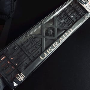 Puede incluir: Un marco negro para matrícula con la palabra "UKRAINE" en letras plateadas. El marco tiene un diseño de rejilla complejo y un emblema plateado. El marco está en un embalaje de plástico transparente.