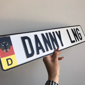 May include: A white and black personalized license plate with the text "DANNY LNG" and a German flag with the letter "D" in the lower right corner.