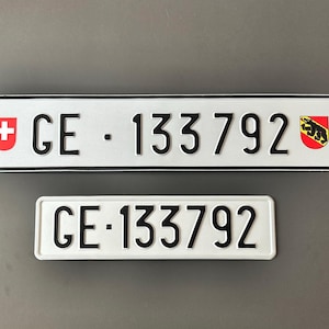 Puede incluir: Dos matrículas blancas con texto y bordes negros. La placa superior tiene el texto "GE - 133792" y un emblema de cruz roja y blanca a la izquierda y un escudo a la derecha. La placa inferior tiene el mismo texto.