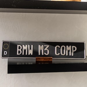 Puede incluir: Placa de matrícula negra con el texto "BMW M3 COMP" en letras plateadas. La placa tiene un símbolo de la Unión Europea y la letra "D" en el lado izquierdo. La placa se exhibe en una estantería negra.
