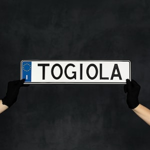 May include: A white license plate with the word "TOGIOLA" in black capital letters. The plate has a blue EU emblem on the left side. The plate is held by two gloved hands against a dark background.