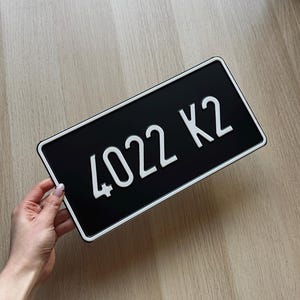 May include: Black rectangular license plate with white text reading "4022 K2". The plate has a white border and is held by a person's hand. The background is a light wood surface.
