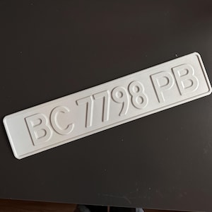 Puede incluir: Placa de matrícula rectangular plateada con el texto en relieve "BC 7798 PB". La placa tiene esquinas redondeadas y está colocada sobre un fondo oscuro.