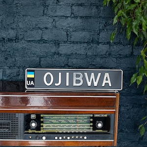 Puede incluir: Una radio de madera vintage con una placa de matrícula gris oscuro que dice "OJIBWA" en letras plateadas. La placa de matrícula tiene una bandera ucraniana y "UA" en el lado izquierdo. La radio está colocada contra una pared de ladrillos azul oscuro.