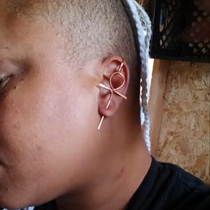 May include: A copper-colored ear cuff featuring a circular design and linear accents. The jewelry piece is worn on the ear, with a minimalist and modern aesthetic. The ear cuff is a unique accessory.