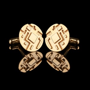 Könnte beinhalten: Ein Paar goldene Manschettenknöpfe mit geometrischem Design. Die Manschettenknöpfe sind rund und haben ein erhabenes Muster.
