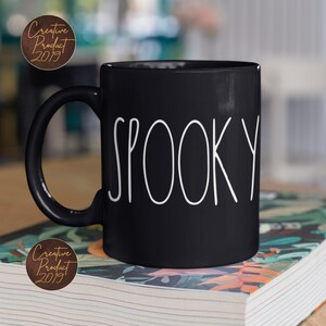 May include: Black ceramic coffee mug with white text that reads "SPOOKY". The mug is sitting on a stack of books with a floral pattern.