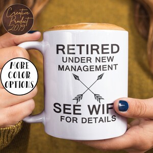 May include: White ceramic coffee mug with black text that reads "Retired under new management see wife for details". The text is accompanied by two crossed arrows.