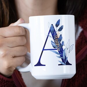 May include: White ceramic mug with a blue and gold floral design and the letter "A" in blue. The mug is being held by a person's hand.