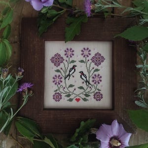 May include: A framed cross-stitch featuring two birds facing each other amidst purple flowers and green foliage. The birds are black and white, and a small red heart is stitched below them. The frame is made of dark brown wood.