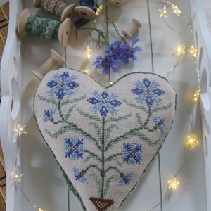 Può includere: Un cuscino a punto ago in forma di cuore con un motivo floreale con fiori blu e foglie verdi. Il cuscino è esposto in un vassoio bianco con luci a catena e bobine di filo.