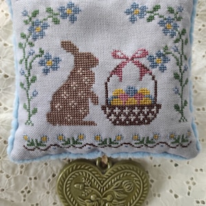 May include: A light blue pillow with a brown bunny and a basket of colorful eggs embroidered on it. The pillow is attached to a green heart-shaped charm.