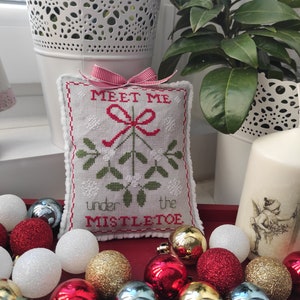 Puede incluir: Una almohada blanca con bordado rojo y verde que representa una rama de muérdago con el texto "Meet me under the mistletoe". La almohada está rodeada de adornos navideños rojos, dorados y blancos.