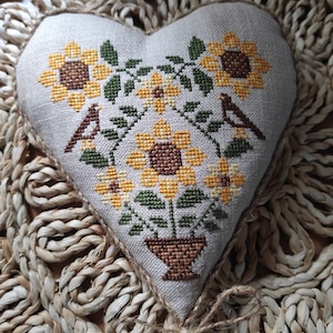 Könnte beinhalten: Ein herzförmiges Kissen mit einem Kreuzstich-Design von Sonnenblumen in einem Topf mit zwei Vögeln. Das Kissen ist aus natürlichem Leinenstoff gefertigt und hat einen rustikalen, handgefertigten Look.