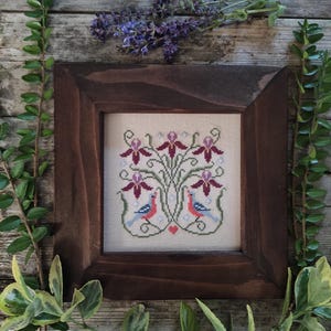 Puede incluir: Un bordado enmarcado con un diseño floral y de aves. El bordado presenta flores de color burdeos, pájaros azules y rojos y un pequeño corazón rojo. El marco es de color marrón oscuro y el fondo es de tela beige claro.