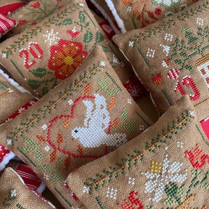 Puede incluir: Un conjunto de 10 adornos navideños hechos a mano, cada uno con un diseño diferente. Los adornos están hechos de tela de arpillera y están decorados con bordados de punto de cruz. Los diseños incluyen una paloma, una flor y un copo de nieve. Los adornos están numerados del 1 al 10.