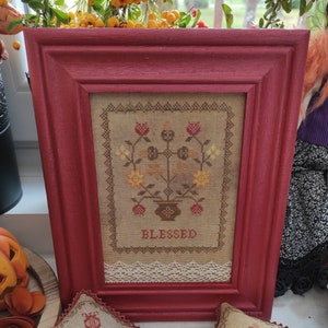 May include: A framed cross-stitch with a floral design and the word "BLESSED" in brown lettering. The frame is a deep red color and has a white lace trim.