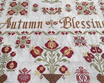 May include: Cross-stitch embroidery on linen fabric with a floral design and the text "Autumn Blessing". The design features red, green, and brown flowers in a vase.