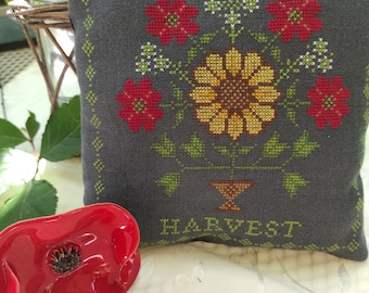 May include: A grey cushion with a cross-stitch design of a bouquet of red and yellow flowers in a brown vase. The cushion has the word "HARVEST" stitched in green below the flowers.