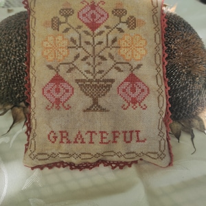 May include: A beige and brown embroidered pillow with a floral design and the word "GRATEFUL" stitched in red. The pillow has a red crocheted border.