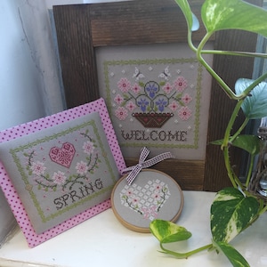 Peut inclure: Trois broderies au point de croix encadrées sur le thème floral rose et blanc. La pièce du haut dit "Bienvenue", la pièce du milieu dit "Printemps" et la pièce du bas est un cœur avec des fleurs.