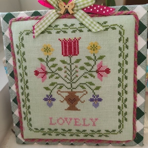 Pode incluir: Um bordado de ponto cruz verde com um desenho floral que apresenta flores rosa, vermelhas, amarelas e roxas em um vaso marrom. O bordado é emoldurado com uma borda xadrez verde e branca e tem a palavra "LOVELY" bordada em rosa abaixo das flores.