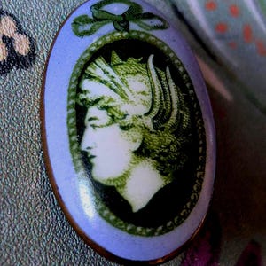 Puede incluir: Broche ovalado con borde azul claro y retrato detallado de una mujer de perfil. El retrato es en tonos verdes y blancos, con una corona de laurel y un lazo. El broche tiene un borde color cobre.